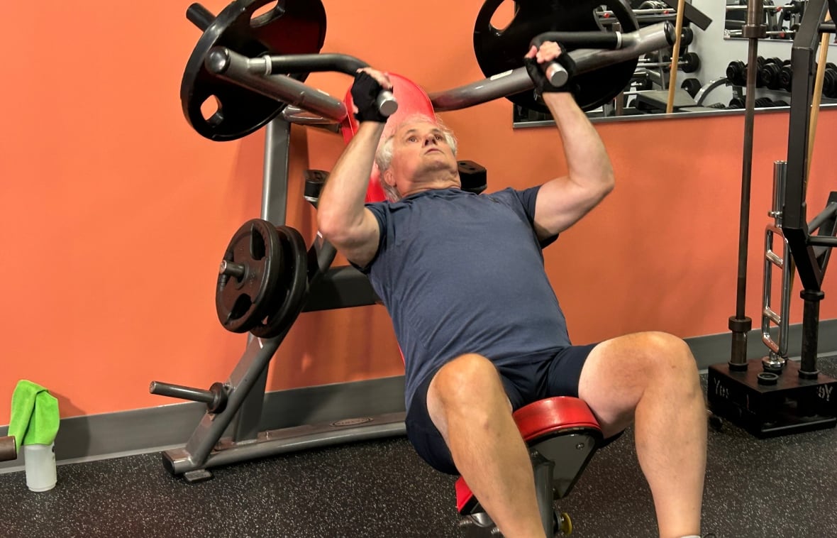 A white man with white hair, wearing blue t-shirt and black shorts doing shoulder presses at a gym.