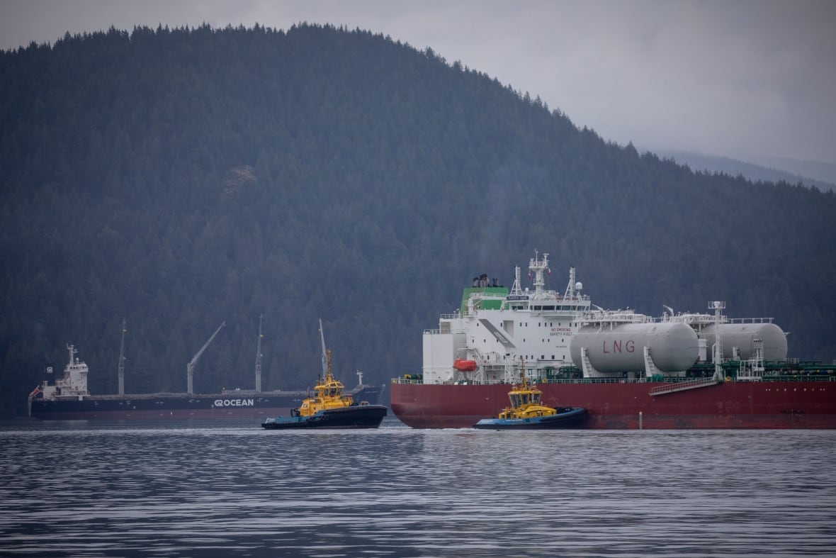 An oil tanker is seen next to two smaller tug boats on a water body, with mountains in the background.