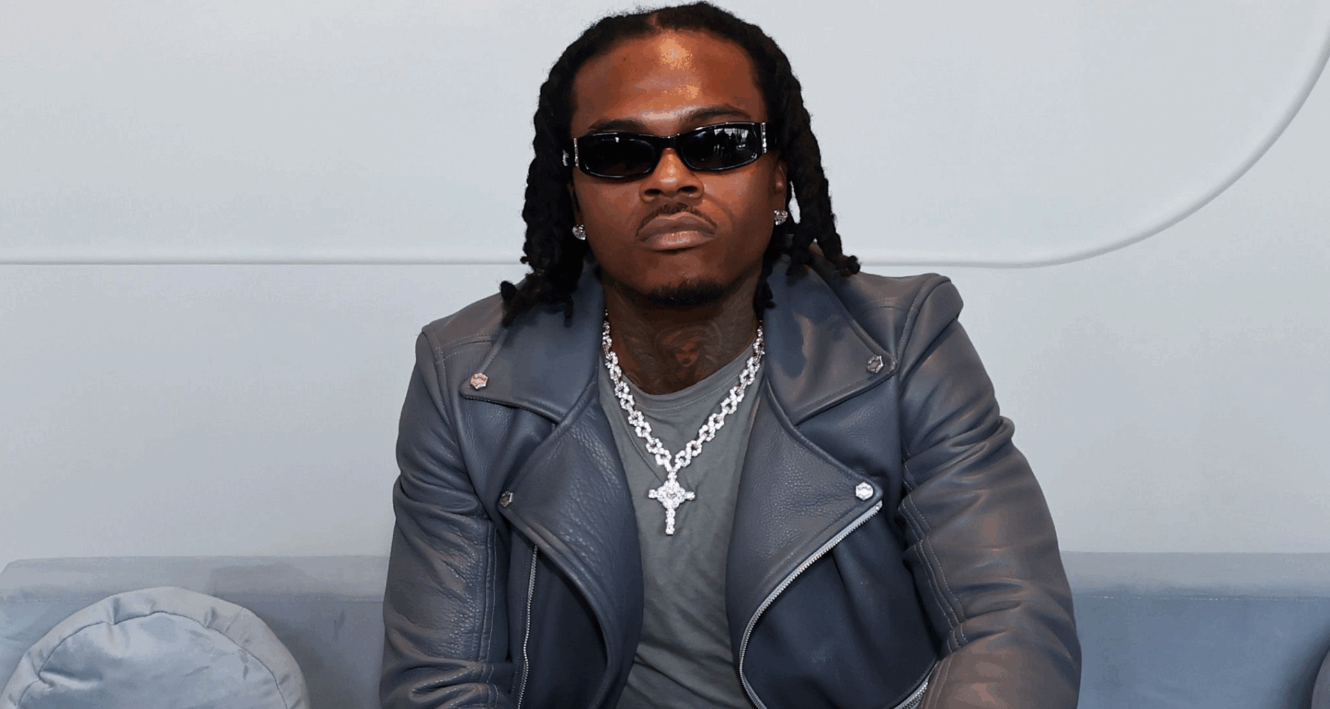 A man wearing sunglasses and a leather jacket sits on a couch, looking at the camera.