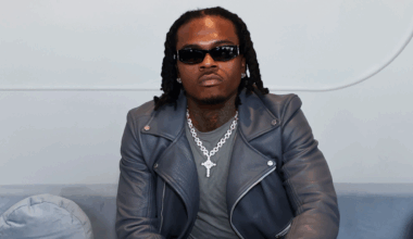 A man wearing sunglasses and a leather jacket sits on a couch, looking at the camera.