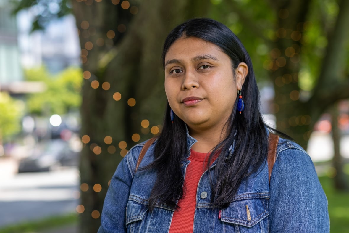 Stacey Gomez has long, black hair and is wearing a blue denim jacket. She is looking at the camera with lights and trees in the background.