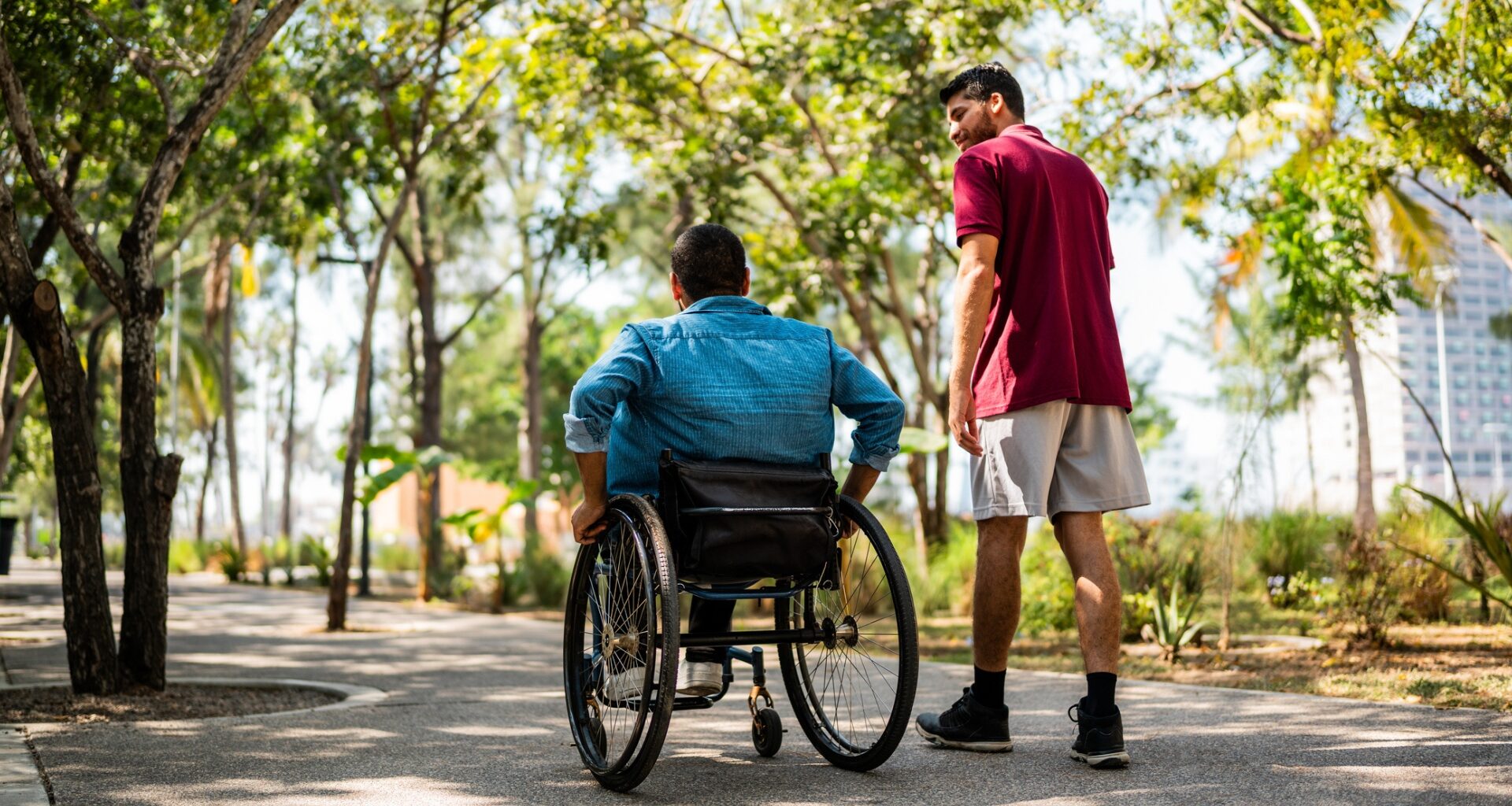 man walking outside with someone in a wheelchair on a path with trees all around in a park setting.