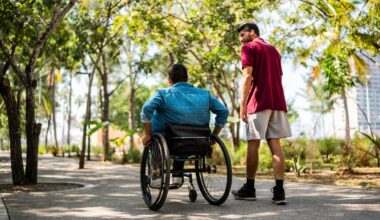 man walking outside with someone in a wheelchair on a path with trees all around in a park setting.