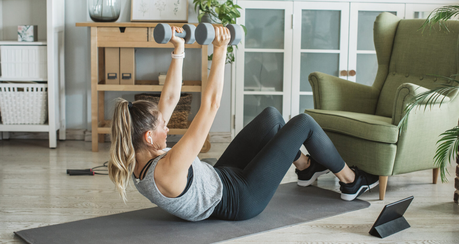 Woman performs sit-ups with dumbbells at home, with her feet hooked under a green armchair