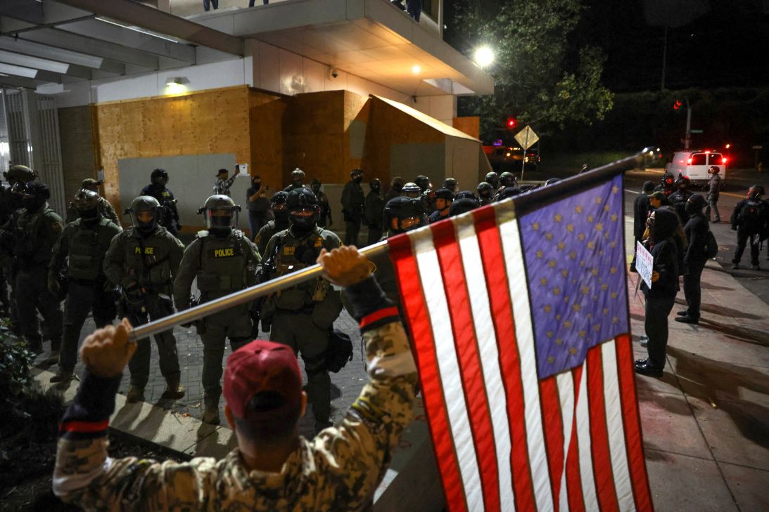 A man holds an American flag as law enforcement officers guard the entrance to an Immigration and Customs Enforcement building in south Portland, Oregon, on October 3, 2025.