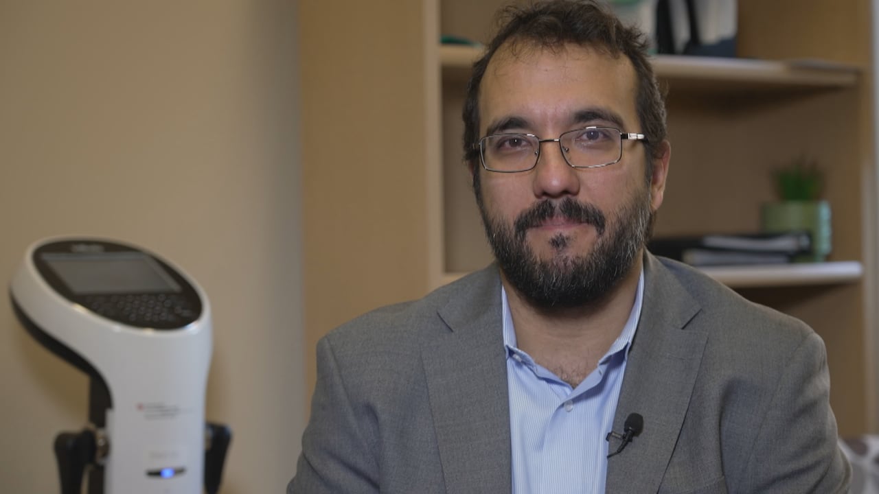 A man wearing a blue shirt smiles at the camera. There is a scale and books behind him.