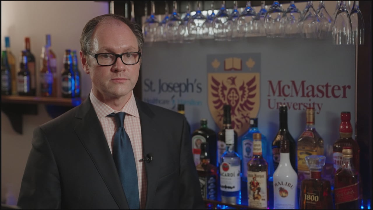 A man stands in front of bottles of alcohol and a sign which reads 'McMaster University' and 'St Joseph's Healthcare'. 