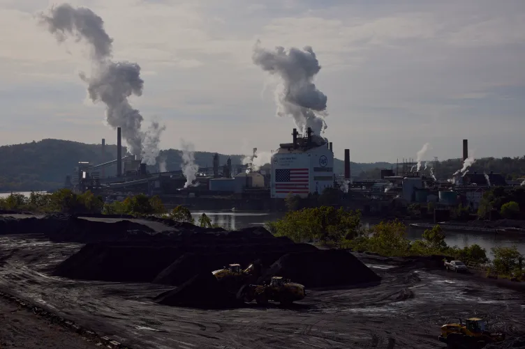 Yellow trucks navigate large piles of black sandy coal in front of a river. Across the river is a large industrial complex with thick steam billowing from several buildings, one of which has an American flag painted on the side. 