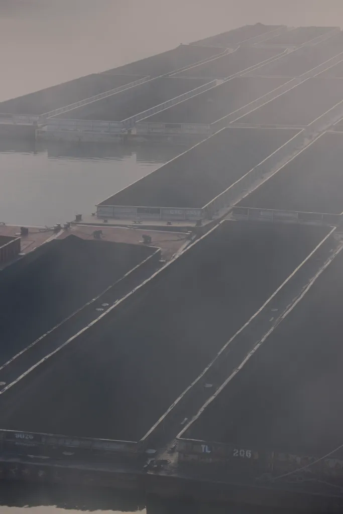 An aerial view of large rectangular barges carrying black sandy material.