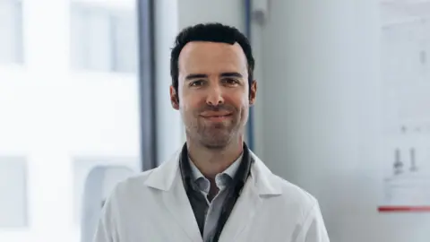 Verley Stéphane Mac Millan wearing a white lab coat and smiling into the camera.