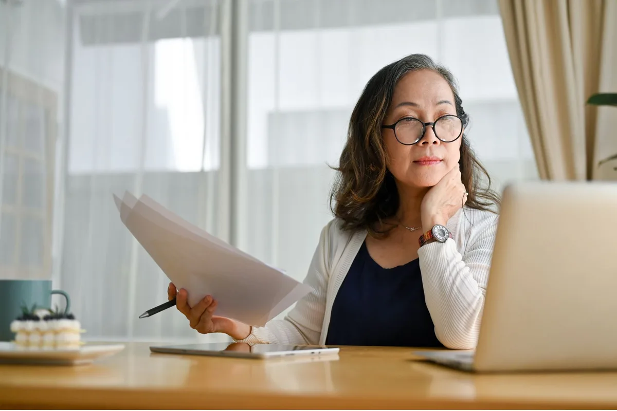 A woman looking at a laptop at her desk, while holding papers