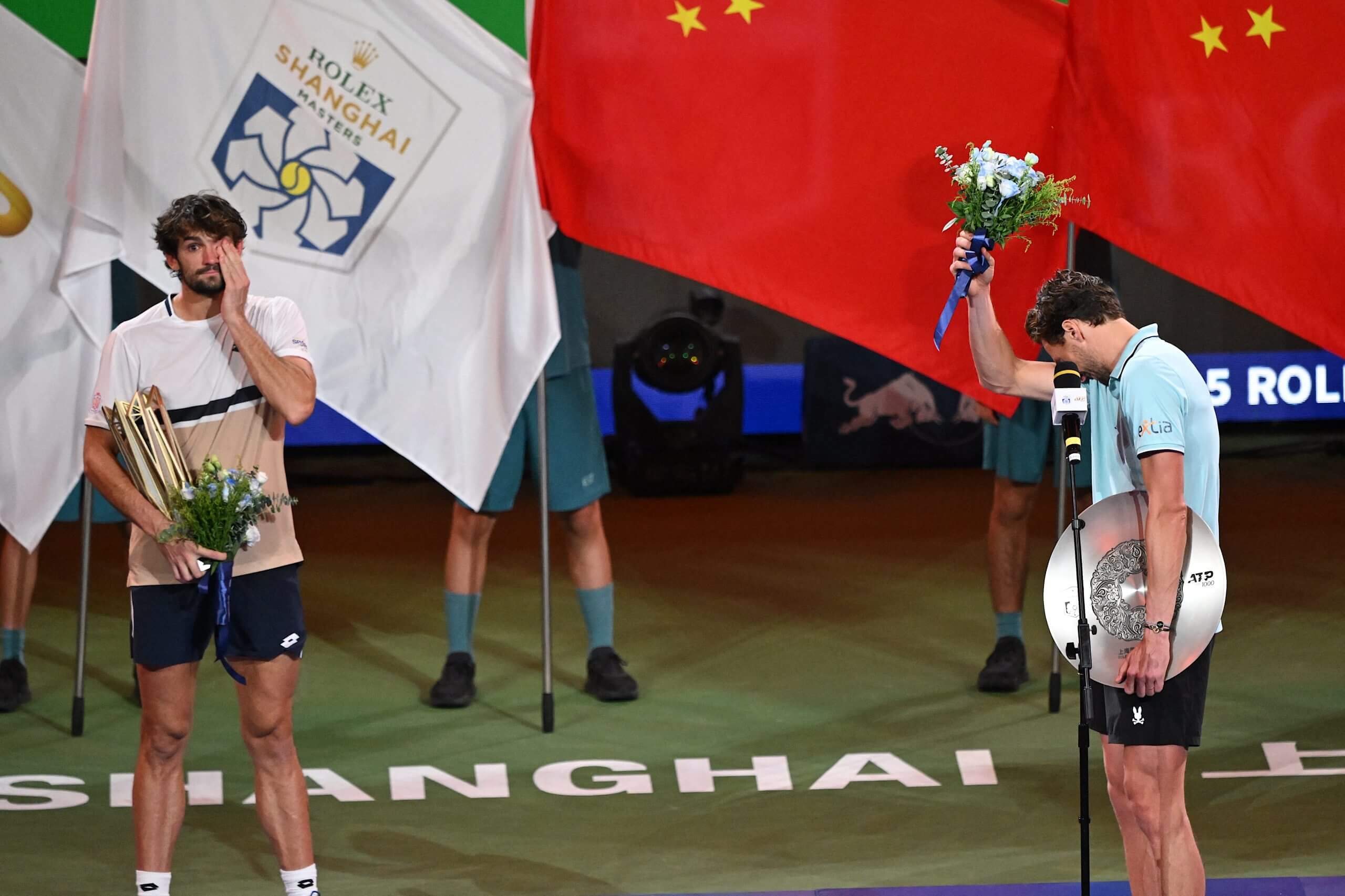 Valentin Vacherot (left) and Arthur Rinderknech (right) stand on a stage holding trophies and flowers, wiping away tears from their eyes.