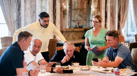 Carin Thakrar / Arora Group Four men sitting around a table with one man standing behind and leaning over and a woman standing and holding a large notepad. There are glasses and cups on the table.