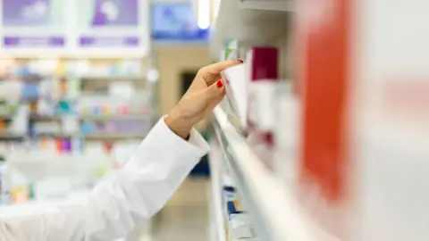 Getty Images Woman takes a box of medicine from a shelf lined with boxes of medicine. She has red nail varnish on her nails.