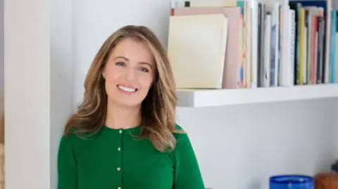 Serena Bolton Smiling into the camera and wearing a green top, Federica Amati stands next to a book shelf.