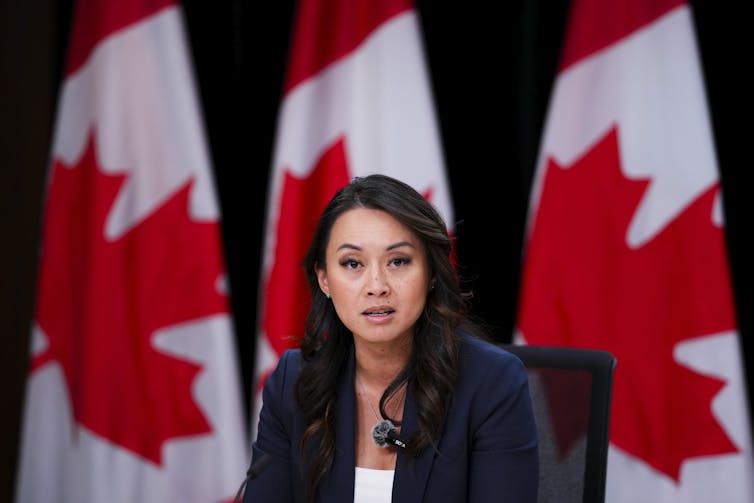 A Filipino woman with long, dark hair in a blazer speaks to someone off-screen while sitting in front of a row of Canadian flags
