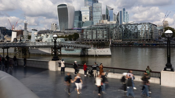 The City of London skyline with modern skyscrapers, the HMS Belfast on the river Thames and people walking along the riverside.