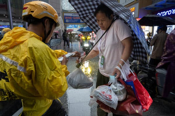 A man in a yellow raincoat and bike helmet talks to a woman holding several plastic bags and an umbrella.