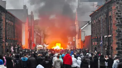 Pacemaker A group of people standing on a street with black stoned houses. There is a police jeep on fire in the background.