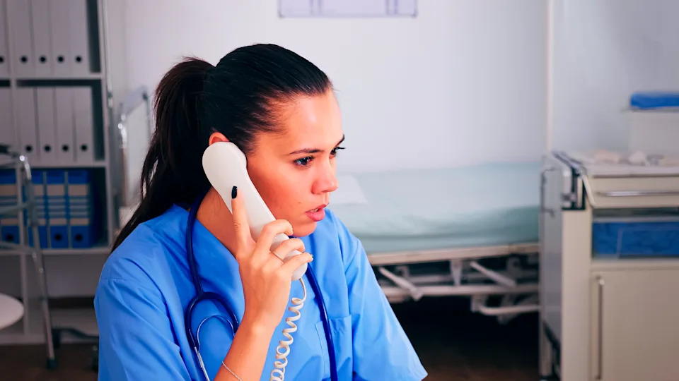 A healthcare worker in scrubs is on the phone in a medical setting, appearing attentive. A hospital bed and medical equipment are visible