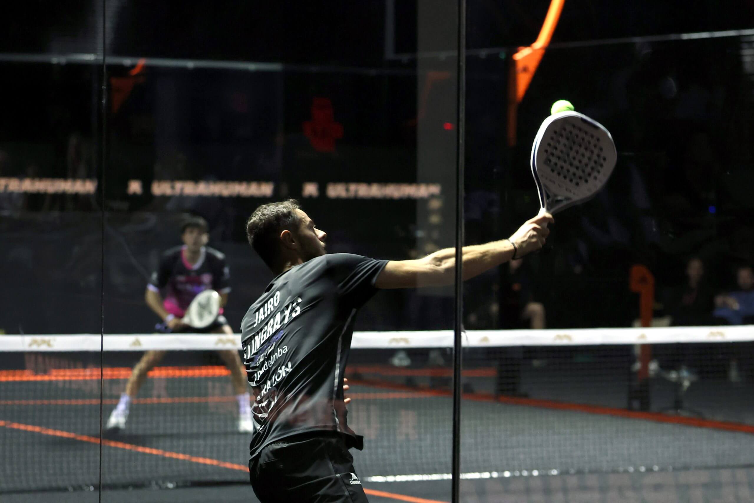 A man reaches up to his right to strike a ball on a padel court, with his opponent waiting in the ready position across from him.