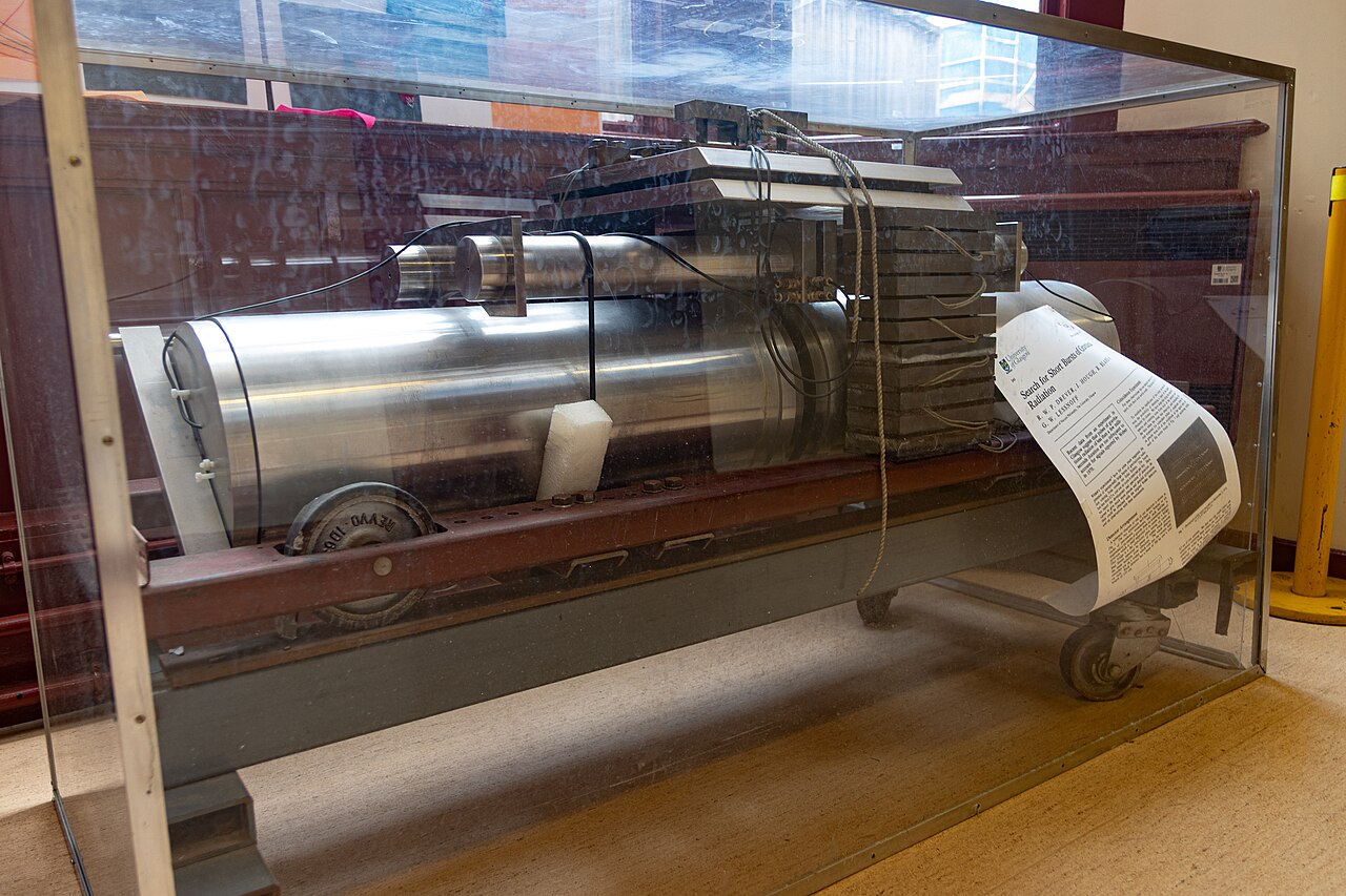 A large, metallic mechanical device—resembling those used to detect Weber bars and gravitational waves—is displayed in a glass case with an informational sheet attached. The industrial machine is set indoors on a tile floor.