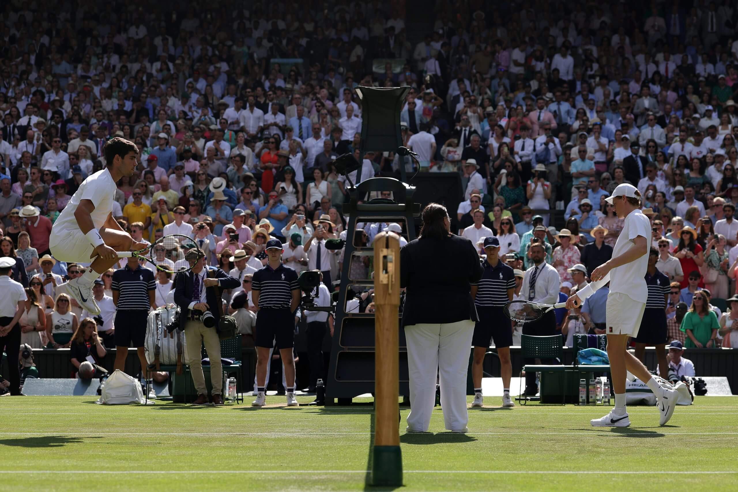 Carlos Alcaraz jumps one side of a tennis net on a grass court as Jannik Sinner shadow-swings on the other side, ahead of the Wimbledon final.