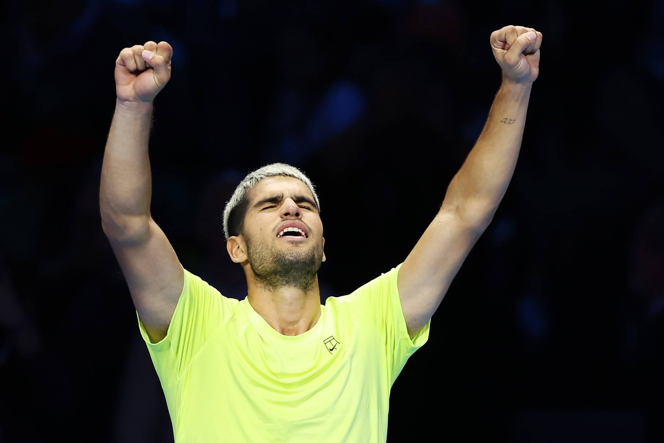 Carlos Alcaraz raises his arms in celebration and closes his eyes in relief, wearing a yellow tennis T shirt.