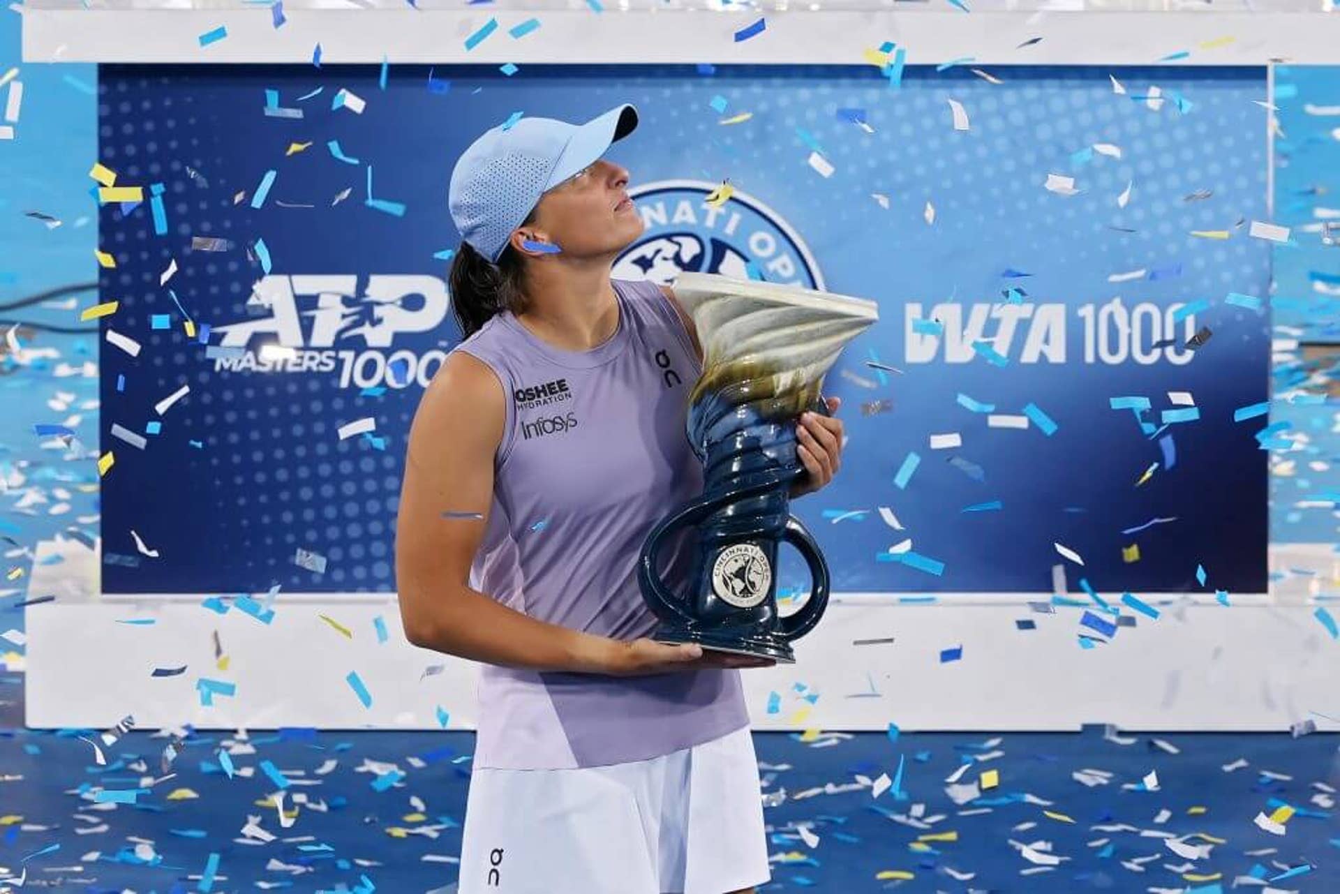 Iga Świątek holds a blue and silver trophy in front of a blue plinth as confetti falls around her. She is looking up and off to her left.