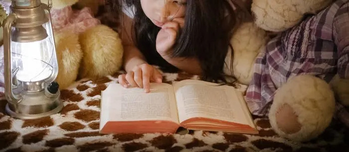 Child reads a book under a makeshift fort with two teddy bears and a lantern