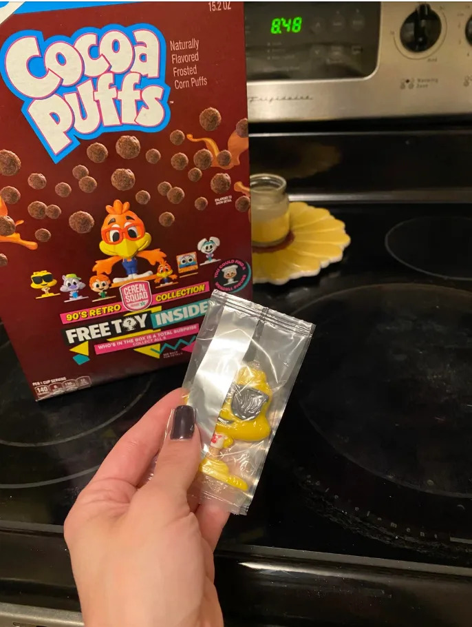 Cereal box of Cocoa Puffs on a stove, with a hand holding a packaged toy from a 90's retro collection