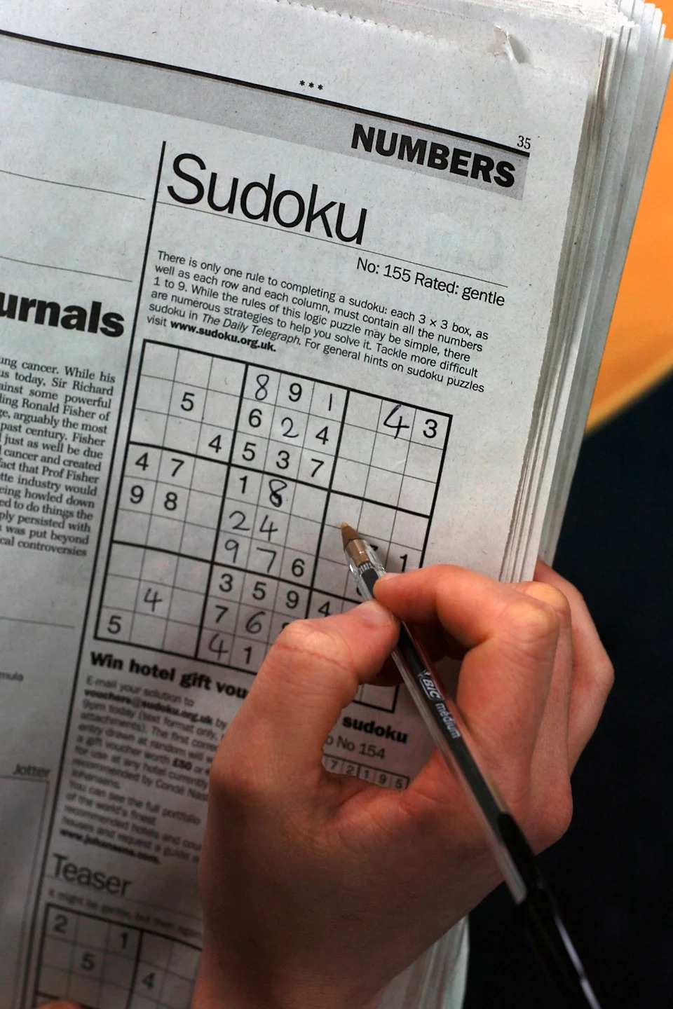 A person fills out a Sudoku puzzle in a newspaper, using a pencil to write numbers in the grid