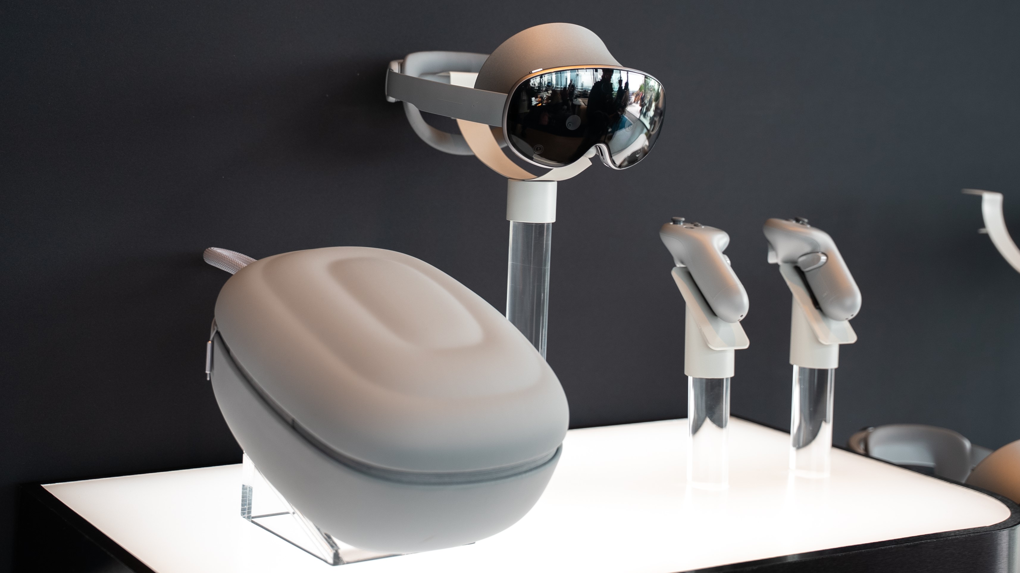 The Samsung Galaxy XR headset, carrying case, and controllers on four pedestals above a white, lit surface.