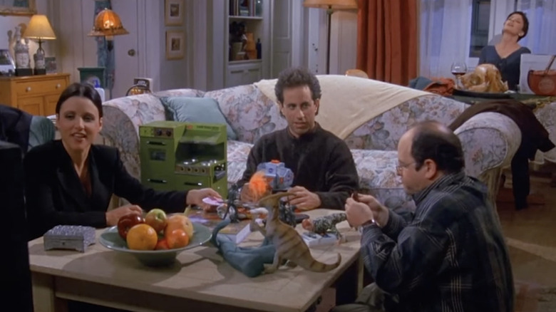 Julia Louis-Dreyfus, Jerry Seinfeld, and Jason Alexander as Elaine, Jerry, and George playing with toys while Julia Pennington as Celia sits unconscious in the kitchen in Seinfeld