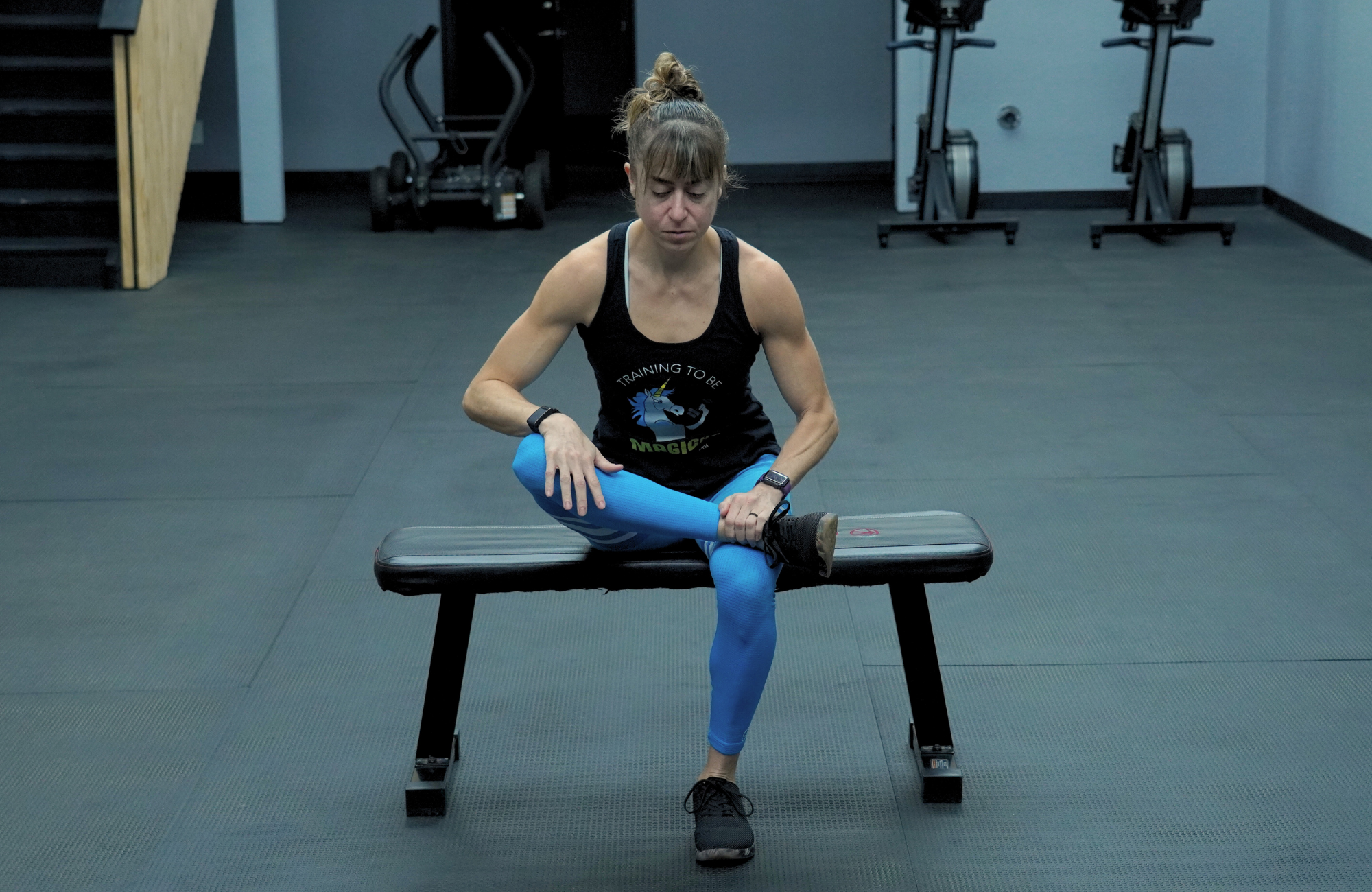 Cori Lefkowith performs a seated pigeon. She is sitting on a weights bench, with her right ankle crossed over her left knee, gently pushing down on her elevated right knee.