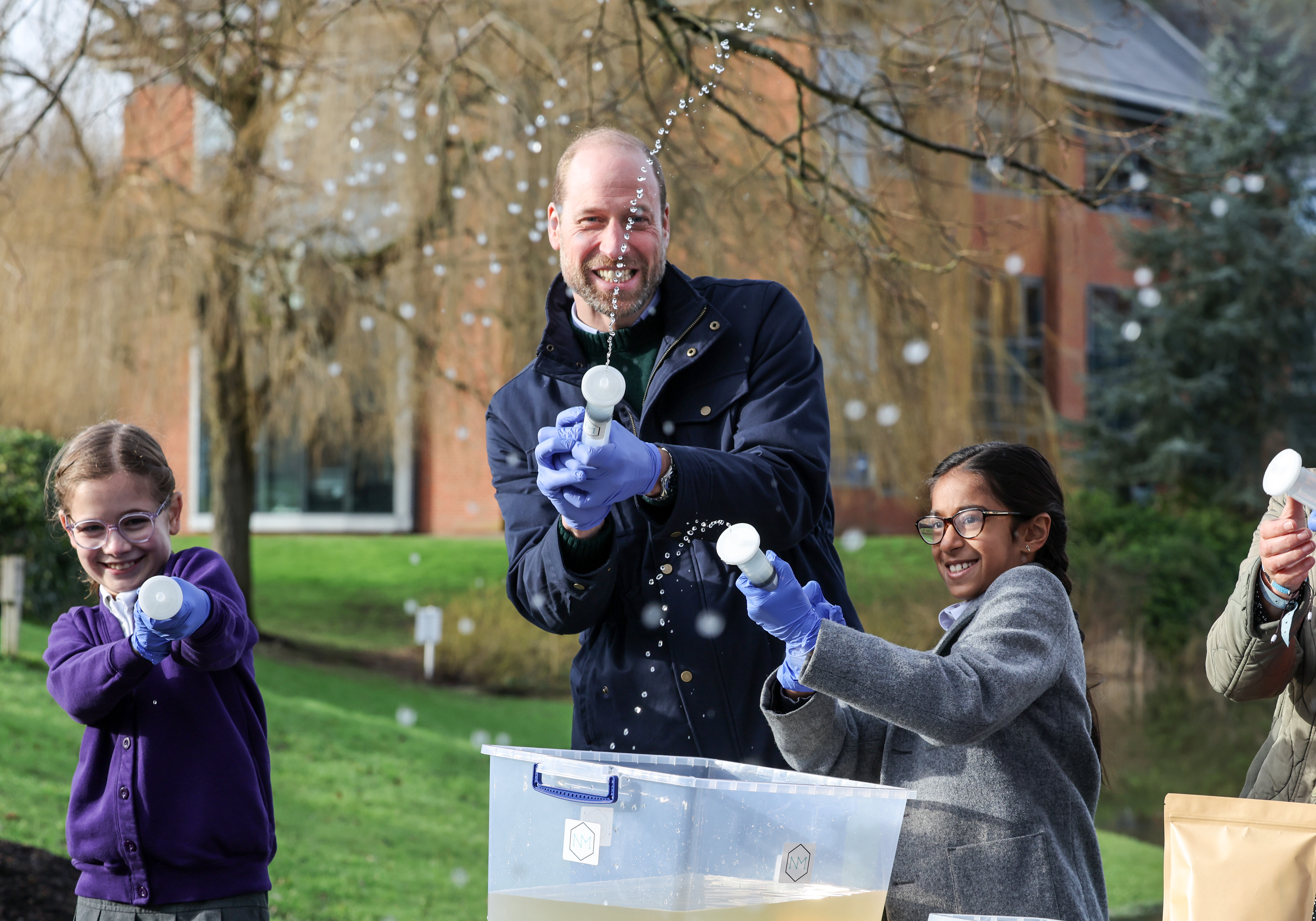 Prince William and two children spraying water from syringes, all wearing blue gloves.