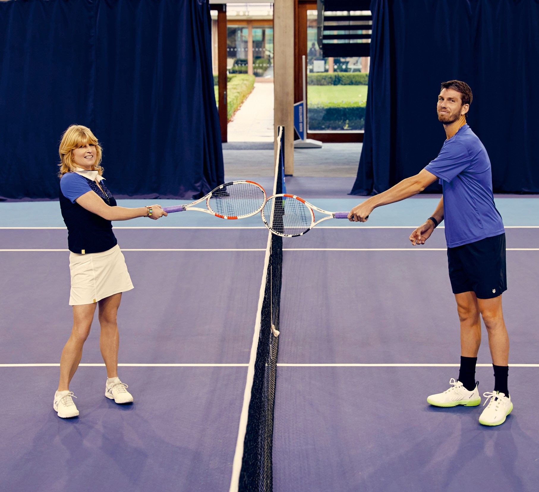Two tennis players, a woman and a man, extend rackets towards each other over the net on an indoor court.
