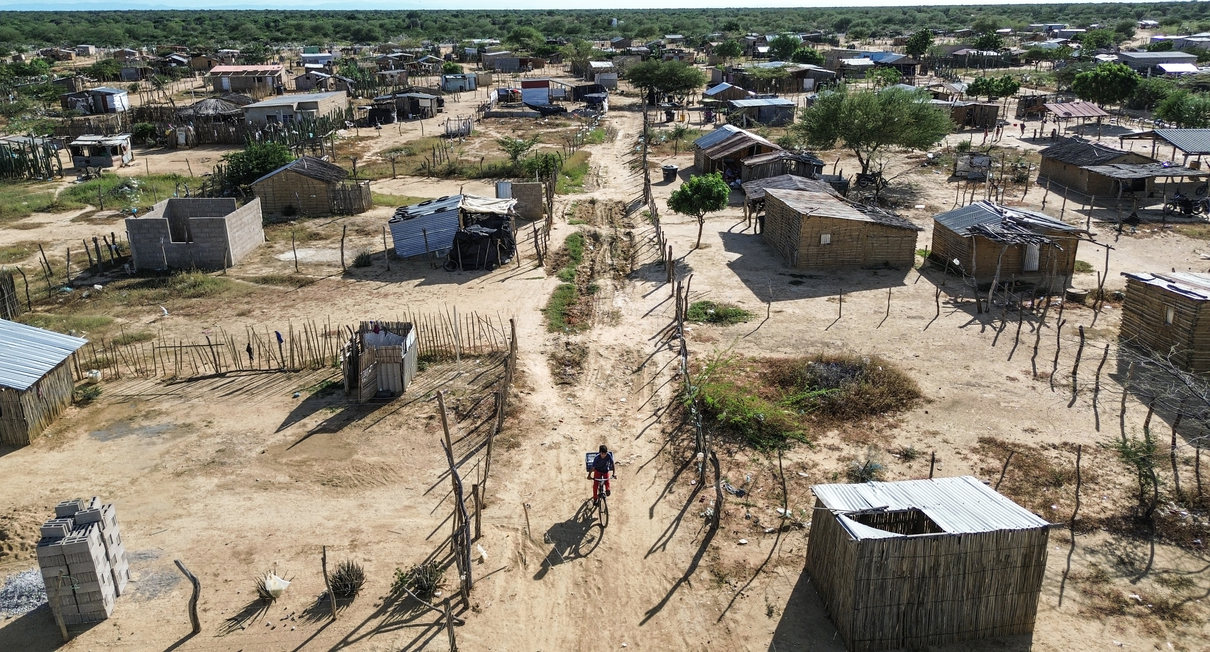 Aerial view of a boy on a red bicycle carrying a white box down a dirt path in a poor rural village.