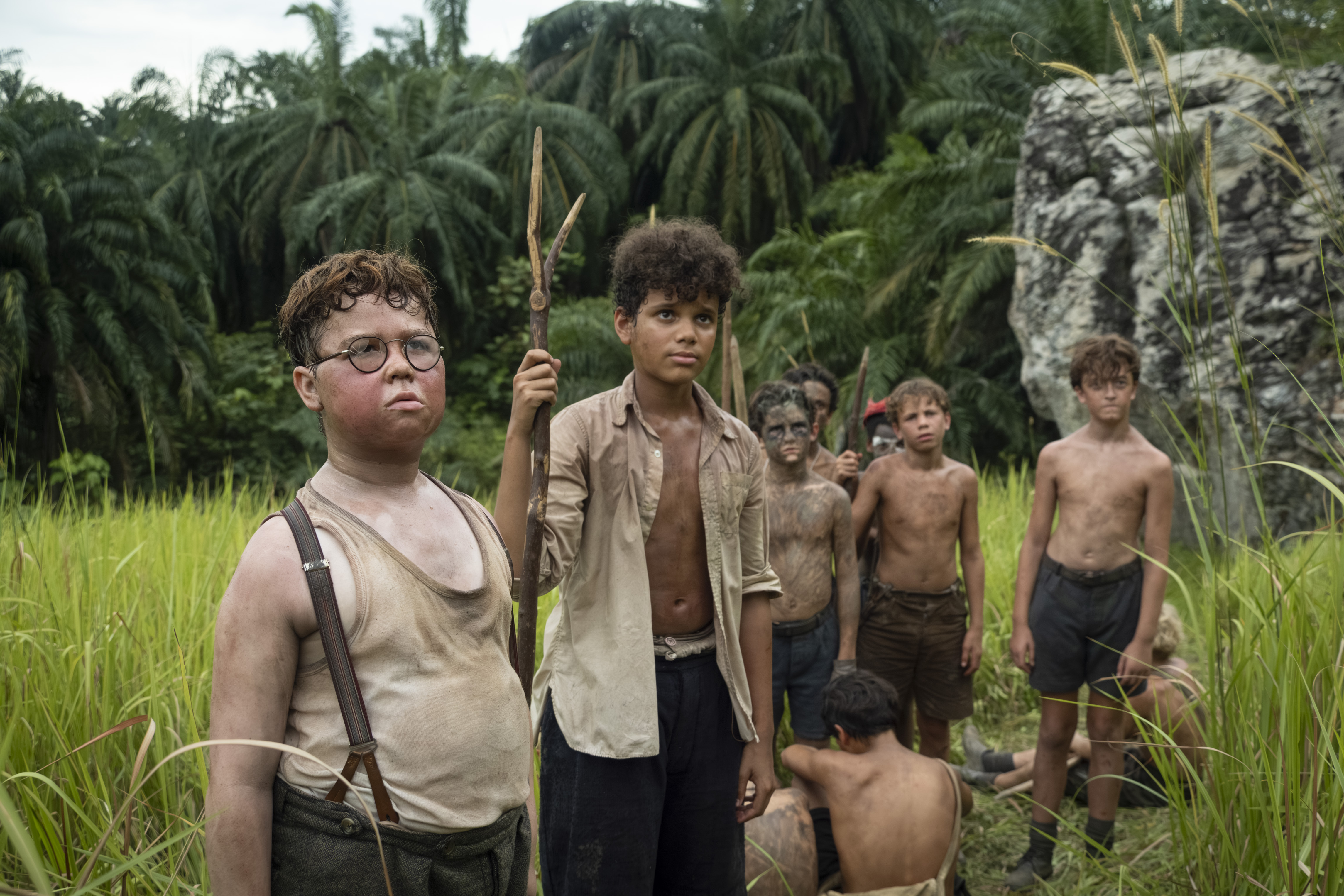 Piggy and Ralph stand in front of Simon and other boys from Lord of the Flies.