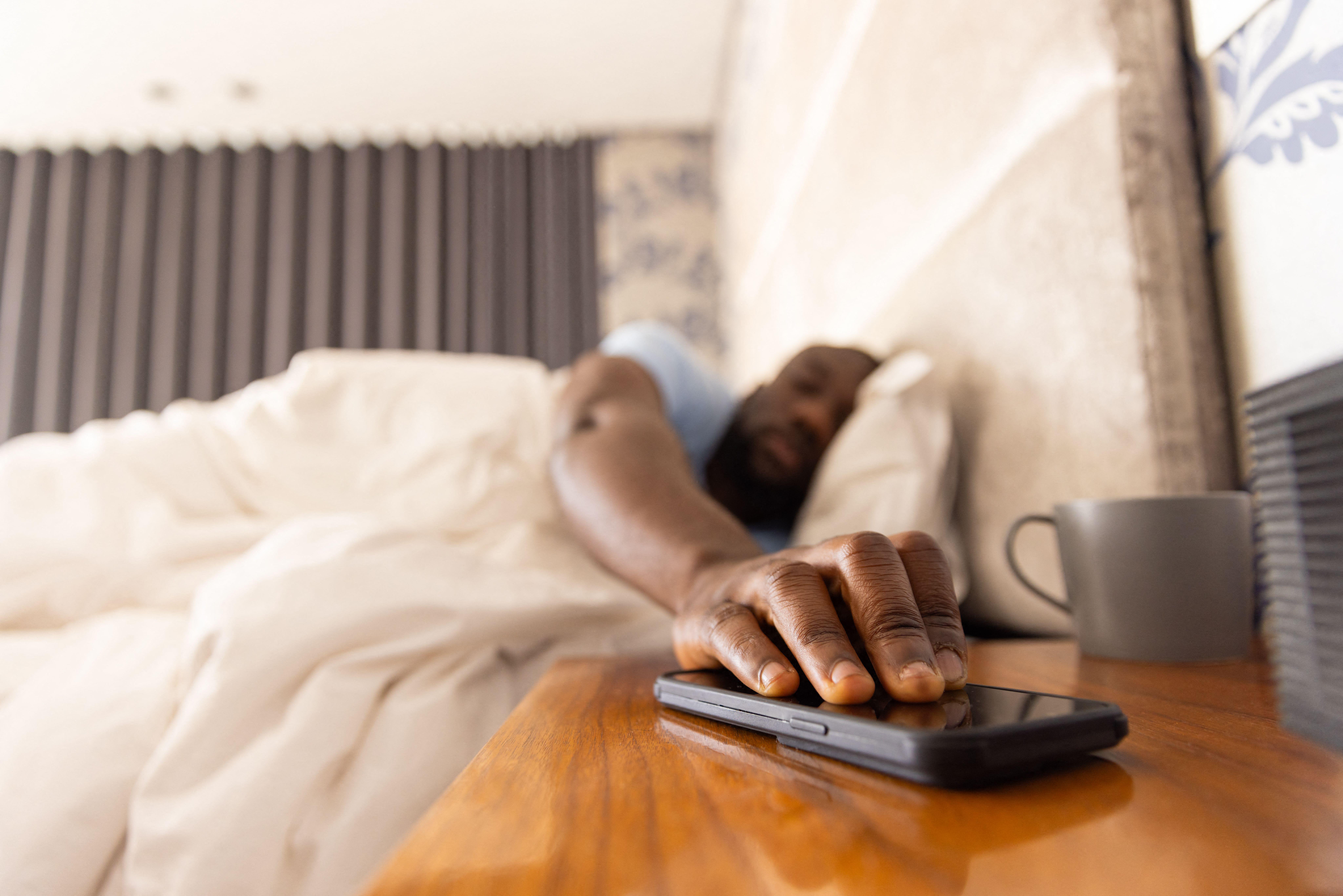 Reaching for smartphone, man waking up in bed with coffee cup on bedside table, copy space