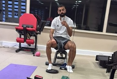 Mo Salah taking a selfie in a gym with various fitness equipment.