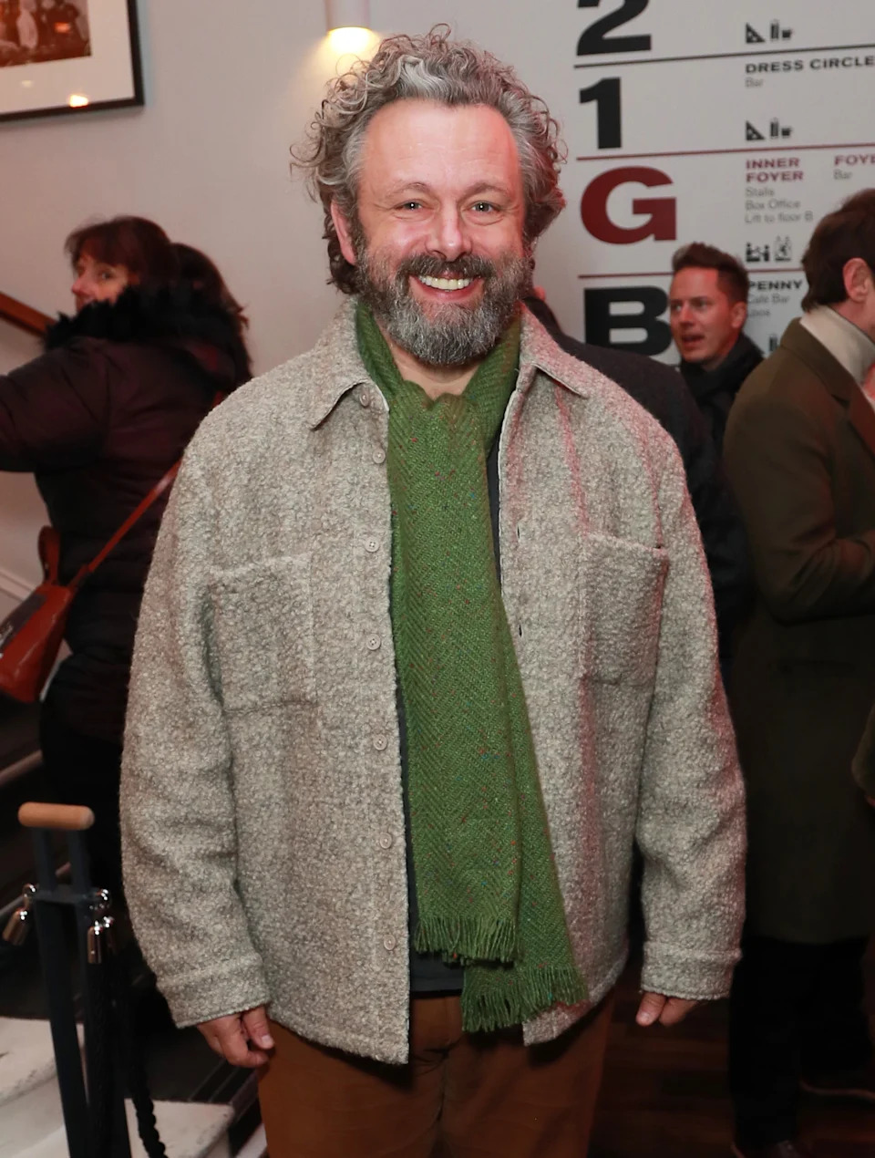 Person in a casual jacket and green scarf smiles in a social setting indoors. People in the background are engaged in conversation