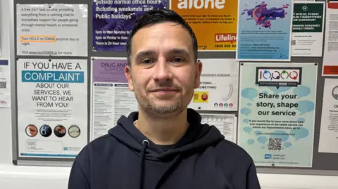 BBC Michael Main is wearing a navy hoodie. He has short black hair and grey facial hair. .He is standing in front of a notice board with lots of coloured posters on it.