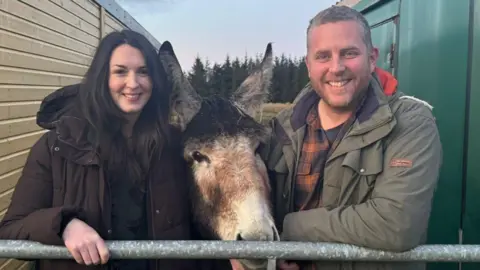 Macaulay College A smiling couple standing a next to a gate with a horse standing between them.