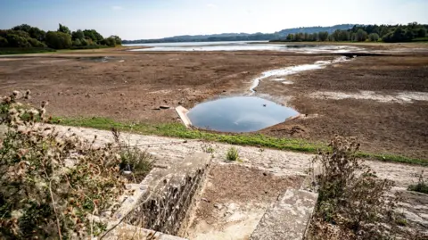 PA Media The exposed lake bed at Chew Valley reservoir. It is a vast open plain covered in mud, with a small pool in the centre. In the foreground there is some bracken and stone steps leading down to where the water level usually is. In the far distance, there is shallow water in the deeper areas of the reservoir.