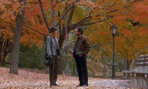 Two people stand talking on a leaf-covered path in a park during autumn, surrounded by trees with fall foliage. One person wears a hat and coat