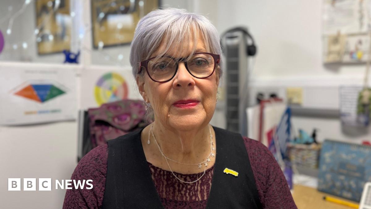 A close-up image of Cherry Cantrell sitting at the desk in her office. She is an older woman, with a purple hue in her short hair. She is wearing  apair of circular glasses, a silver necklace, as well as a purple lace top and black waistcoat. She has a yellow 'Aware NI' pin on her lapel. She is looking at the camera and smiling. Behind her are posters and lights on the wall of her office, but they are blurred.