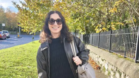 Mary McCarthy A woman with brown wavy hair wearing black sunglasses, a black top and a brown leather jacket smiling. 