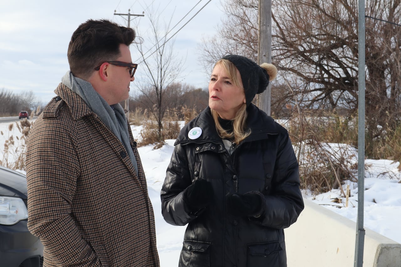 a man wearing glasses, a grey scarf and a winter coat stands beside a blonde woman wearing a black toque, coat and gloves while she speaks.