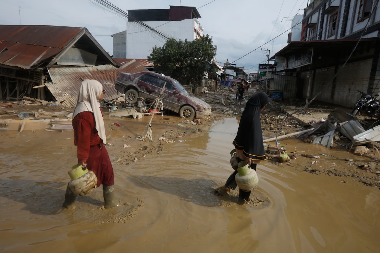 Two people walk down a road flooded with muddy water, carrying objects. 